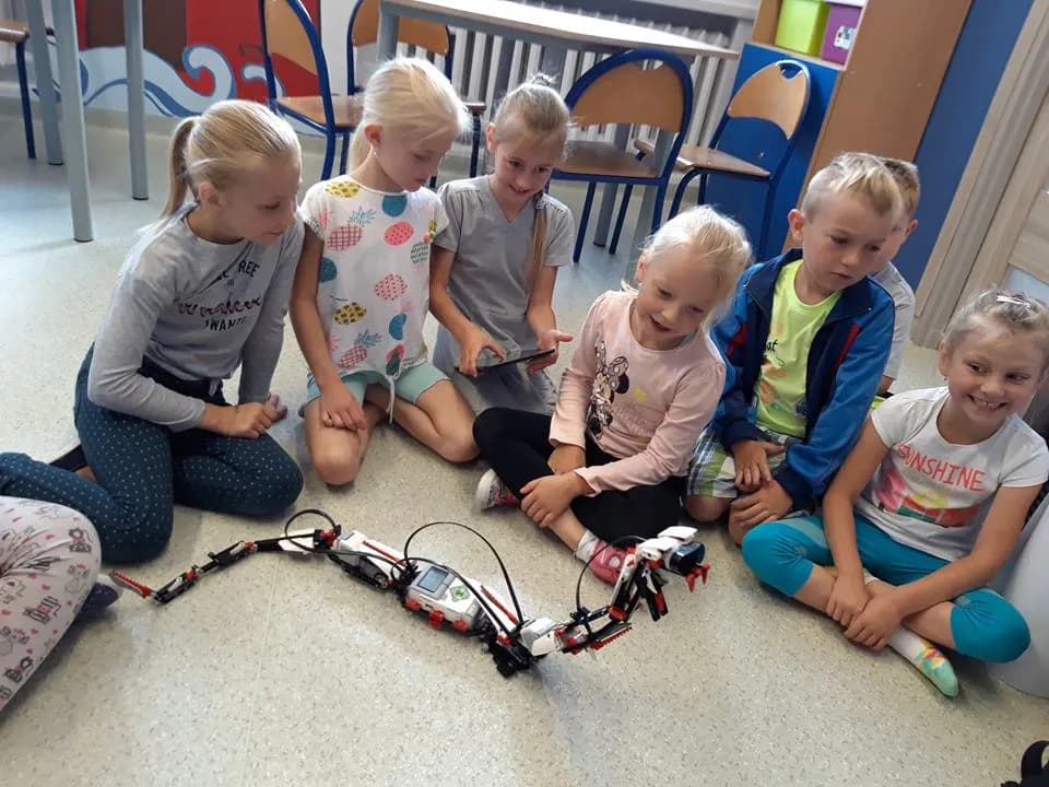 Obraz przedstawia warsztaty z robotyki. Na zdjęciu możemy ujrzeć dzieci zainteresowane robotem przypominającego węża.