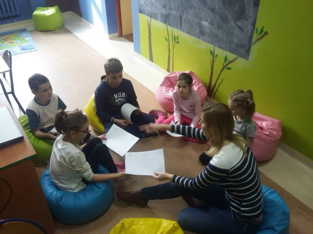 Zdjęcie przedstawia dzieci siedzące w kółku z osobą prowadzącą zajęcia. Zajęcia były o tematyce poczucia własnej wartości.