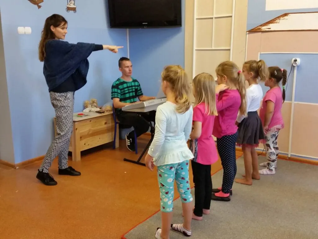 Zdjęcie przedstawia dzieci ustawione w rzędzie w pomieszczeniu z niebieskimi ścianami przygotowujące się wraz z opiekunami do muzycznego występu.