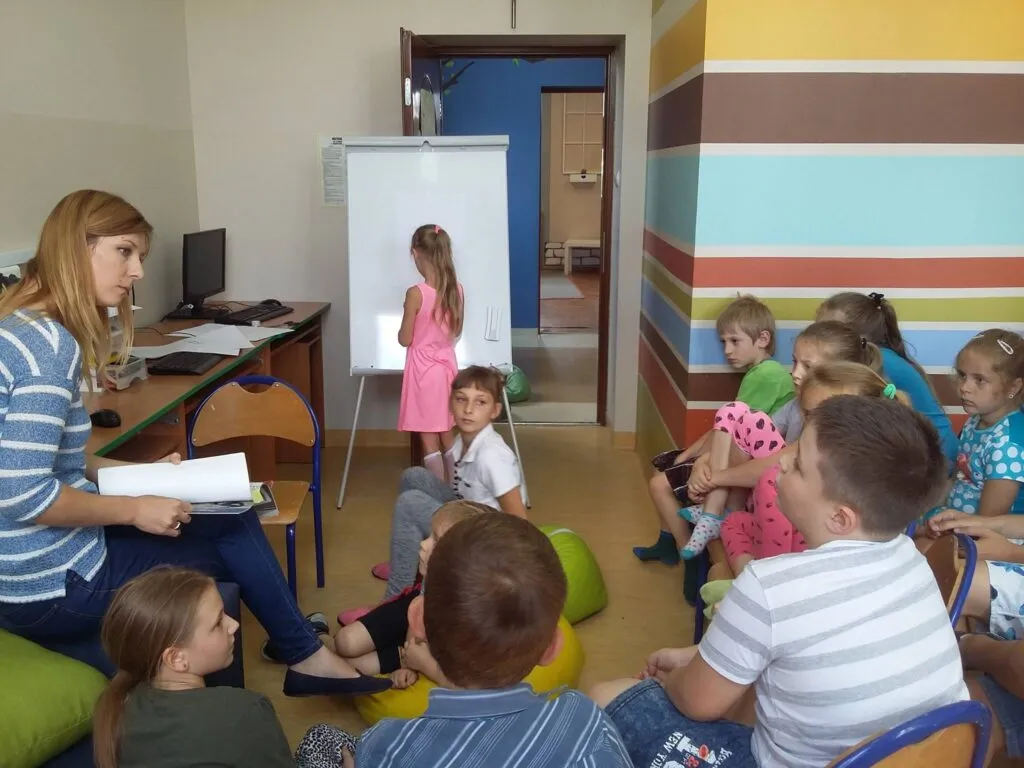 Zdjęcie przedstawia dzieci z placówki oraz osobę prowadzącą zajęcia w pokoju z kolorowymi ścianami. 