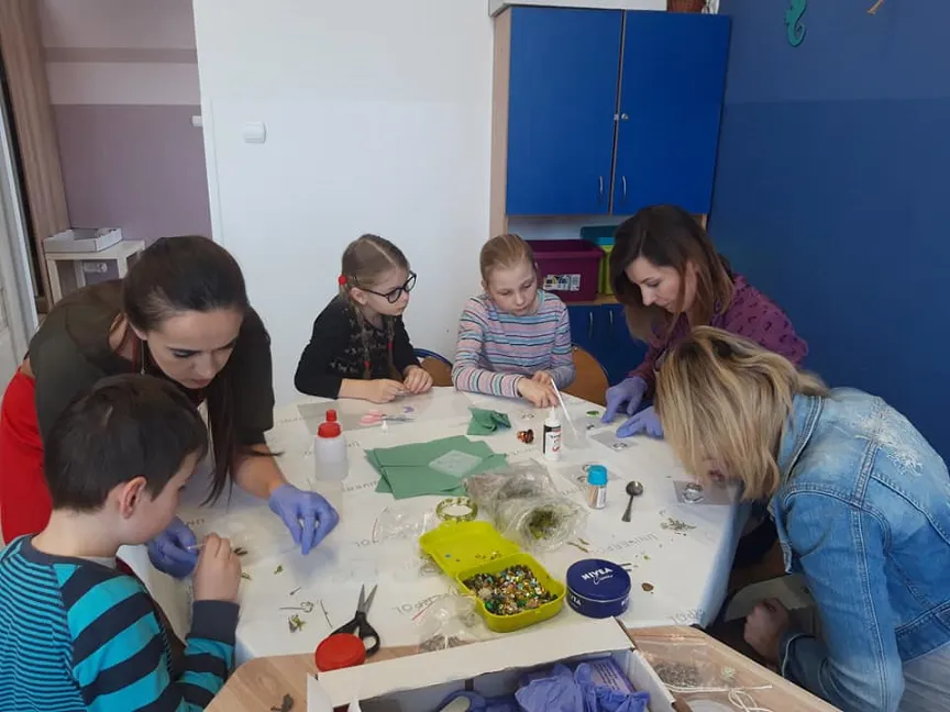 Obraz przedstawia wychowanków SPWD podczas zajęć z wykorzystaniem żywicy epoksydowej. Na zdjęciu możemy ujrzeć dzieci z wychowawczyniami przy stole, na którym powstają przepiękne ozdoby.