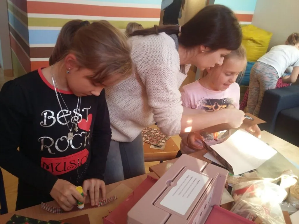 Zdjęcie przedstawia dzieci wraz z osobą prowadzącą zajęcia przy stole tworzące rękodzieła w ramach krainy rozmaitości.