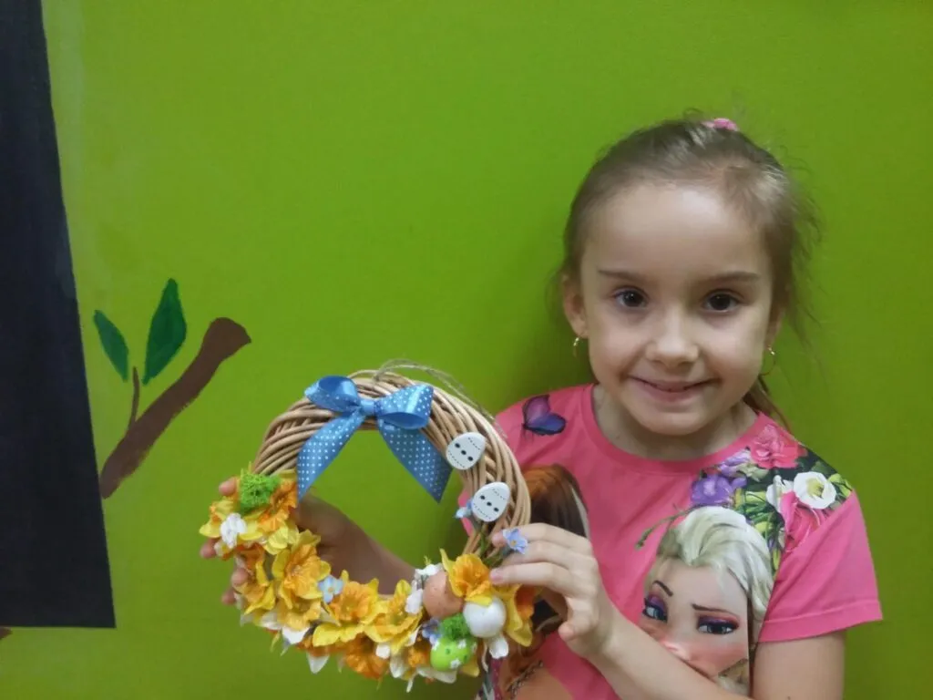 Zdjęcie przedstawia dziewczynkę trzymającą dekorację Wielkanocną na tle zielonej ściany.