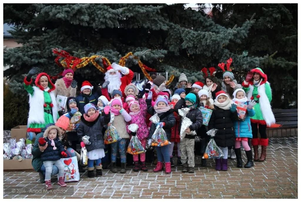 Zdjęcie przedstawia dzieci z placówki na zewnątrz przy dużym drzewie iglastym wraz z św. Mikołajem, który w tym dniu obdarował je prezentami.