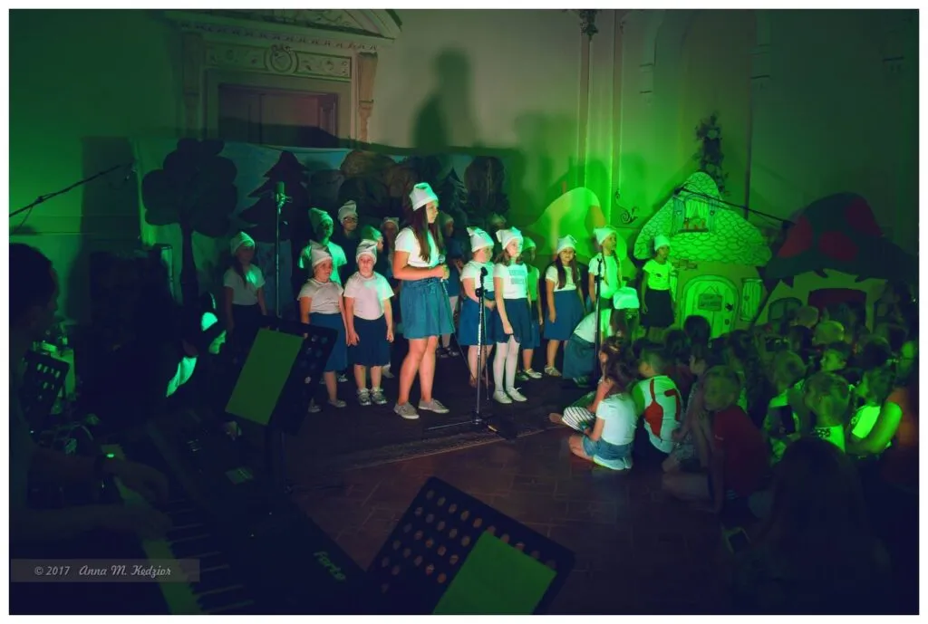 Zdjęcie przedstawia dzieci podczas występu "Kto pod kim dołki kopie" w otoczeniu publiczności i zielonego światła.