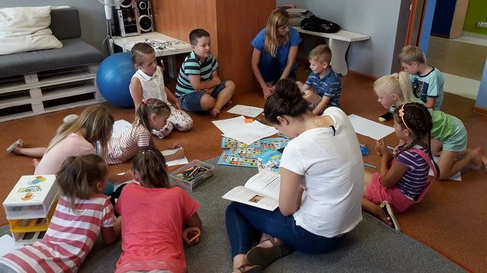 Na zdjęciu widnieje grupka dzieci spędzających czas wraz z osobą opiekująca się nimi. Dzieci na siedzą na podłodze z kartkami oraz czymś do pisania oraz malowania.
