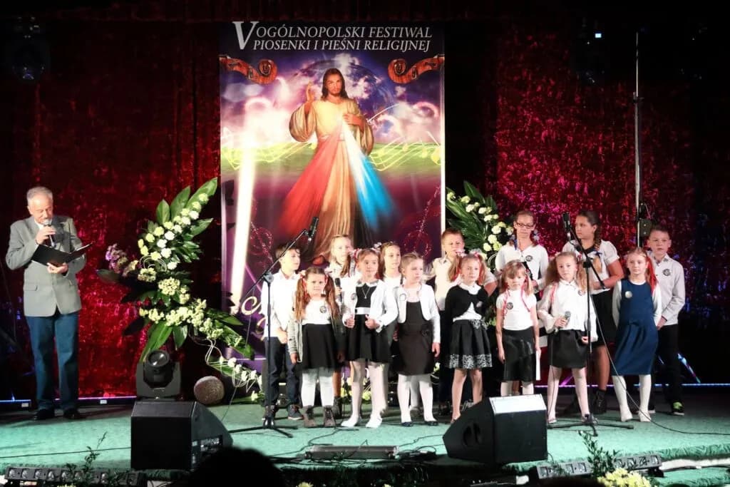 Zdjęcie przedstawia dzieci z placówki na scenie wraz z prowadzącym podczas ogłoszenia wyników konkursu muzycznego. W tle widnieje plakat z Jezusem Miłosiernym.
