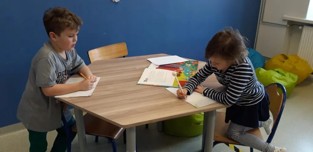 Obraz przedstawia dwójkę dzieci wypełniające karty pracy.