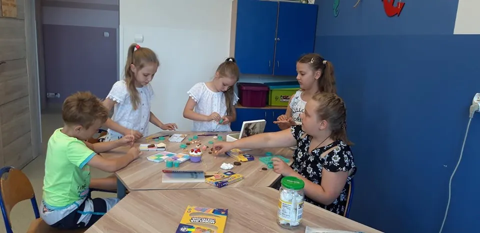 Obraz przedstawia wychowanków SPWD podczas zajęć artystycznych, na których tworzą one różnego rodzaju rękodzieła przy użyciu modeliny czy masy modelarskiej.