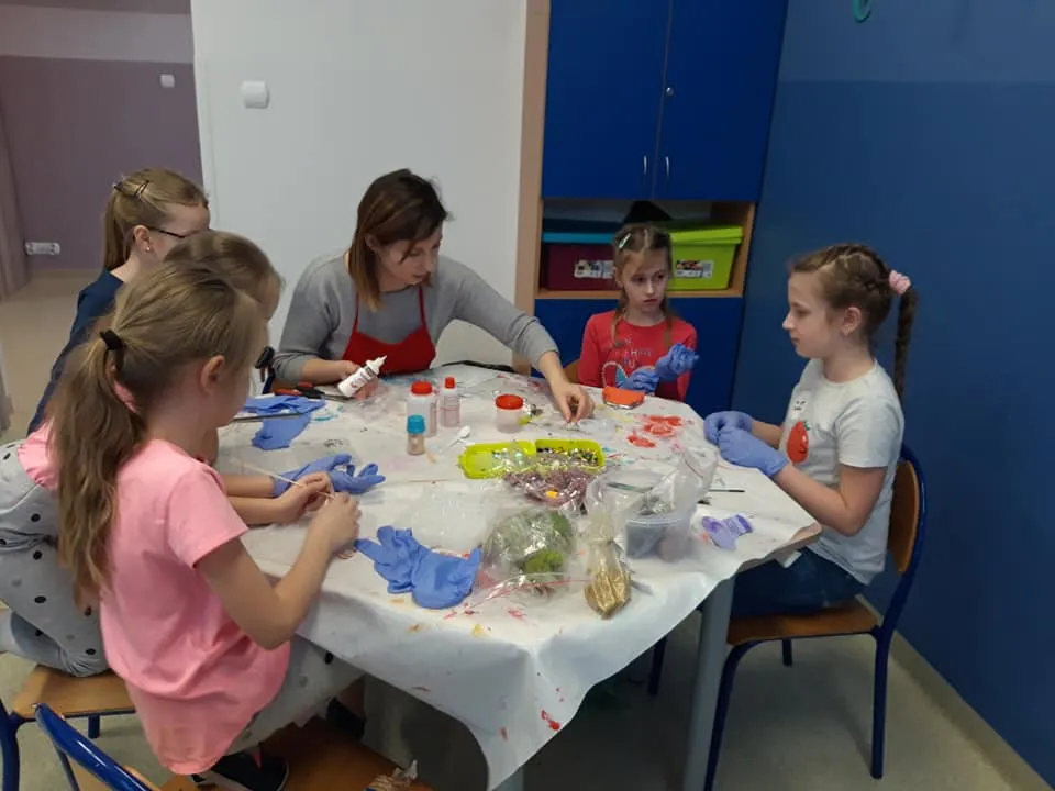 Obraz przedstawia zajęcia artystyczne. Podczas których wychowankowie z wychowawczynią tworzą rękodzieła z żywicy epoksydowej. Na zdjęciu możemy ujrzeć dzieci siedzące przy stole z wychowawczynią oraz różnego rodzaju materiały do tworzenia przepięknych prac.