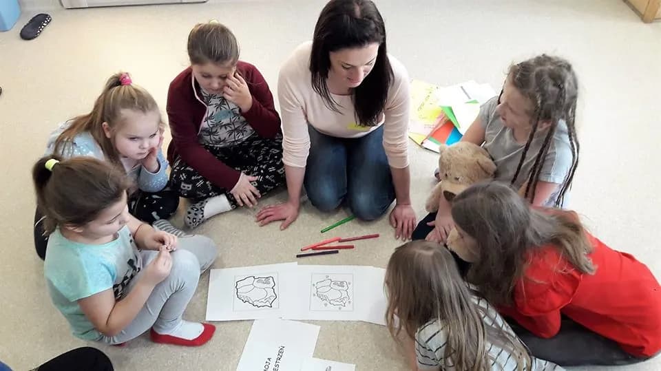 Obraz przedstawia wychowanków podczas zajęć socjoterapeutycznych. Na zdjęciu możemy ujrzeć wychowanków, wychowawczynię oraz kartki na których namalowany jest kontur Polski.